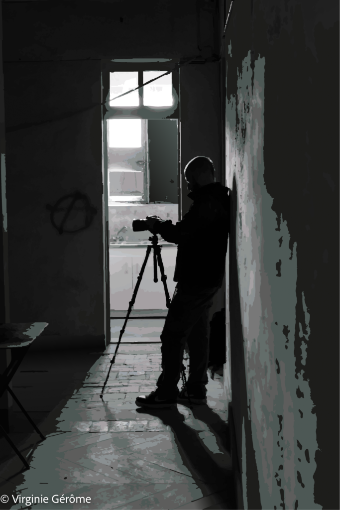 Cette image installe une atmosphère de silence, de retrait et de concentration. Dans un espace intérieur presque entièrement plongé dans l’ombre, une silhouette se détache près d’un mur, occupée auprès d’un appareil posé sur un trépied. Face à elle, une ouverture éclaire le fond de la pièce et devient la principale source de lumière, traçant un contraste net entre obscurité enveloppante et clarté lointaine.

La composition repose sur une tension très forte entre la verticalité du couloir, la présence sombre de la figure humaine et l’appel lumineux de la porte ouverte. La lumière ne révèle pas l’ensemble du lieu : elle découpe, suggère, isole. Les murs usés, les surfaces écaillées et le sol marqué par les reflets deviennent les témoins discrets d’un espace habité par le temps.

La silhouette apparaît presque comme une ombre parmi les ombres. Pourtant, sa posture indique un geste précis, attentif, presque méditatif. L’image semble alors parler autant de l’acte de regarder que de ce qui est regardé. Elle met en scène une présence humaine non pas dans l’exposition directe, mais dans une forme de retrait, comme si voir exigeait ici de se tenir à la frontière entre lumière et obscurité.

Le noir et blanc renforce cette dimension introspective. Il dépouille la scène de tout effet anecdotique pour en faire un espace de perception pure : un lieu où la lumière devient révélation partielle, où l’ombre protège, absorbe et prolonge le mystère.

Dans cette œuvre, « Entre ombres et lumière » prend une dimension presque réflexive : il ne s’agit plus seulement de montrer un lieu ou un passage, mais aussi de suggérer l’acte même d’observer, de cadrer, de chercher une image dans la pénombre.

À ce stade de la série, une nouvelle piste s’ajoute donc au fil possible : après la trace, le passage et l’apparition, il y a aussi le regard — le regard qui cherche, qui attend, qui révèle sans tout montrer.